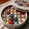 A round box of small, colorful bonbons arranged in separate sections. The bonbons are in shades of red, blue, white, and yellow, neatly organized in distinct sections, each containing 5 to 6 bonbons of the same color. The round box has a floral motif on its lid. The box is open, showing the bonbons inside. The background includes a table or surface where the box is placed.