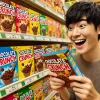 An image showing a person excitedly discovering a variety of similar chocolate crunch products on a store shelf. The person is Japanese and looking happy as they inspect the products, with bright and cheerful colors in the store. The shelf is filled with various colorful packages of chocolate crunch alternatives.