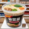 A large plastic container filled with a new release of convenience store ramen, featuring generous portions of noodles, vegetables, and pork. The packaging is new and modern, with eye-catching labels indicating it’s a new product. The container is placed on a promotional display at a convenience store, ready for customers to try.