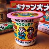 A close-up of a colorful yogurt-flavored candy container similar to 'Morocco Yogurt.' The small plastic cup has a bright and playful design, featuring Japanese characters and a fun illustration. It sits on a wooden table with other traditional Japanese dagashi packaging around, creating a nostalgic atmosphere. The container is neatly sealed with a plastic lid, typical of popular Japanese sweets.