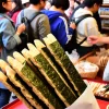 A popular thick, stick-shaped crispy rice cracker wrapped in seaweed, displayed in a traditional Japanese market. The scene shows the snack being highly popular among shoppers, with several people gathered around, browsing and buying the product. The market has a vibrant and lively atmosphere.